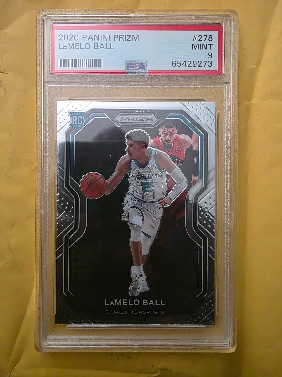 A phone photo of a PSA 9 graded 2020 Panini Prizm LaMelo Ball rookie card, lying on a wooden table in daylight